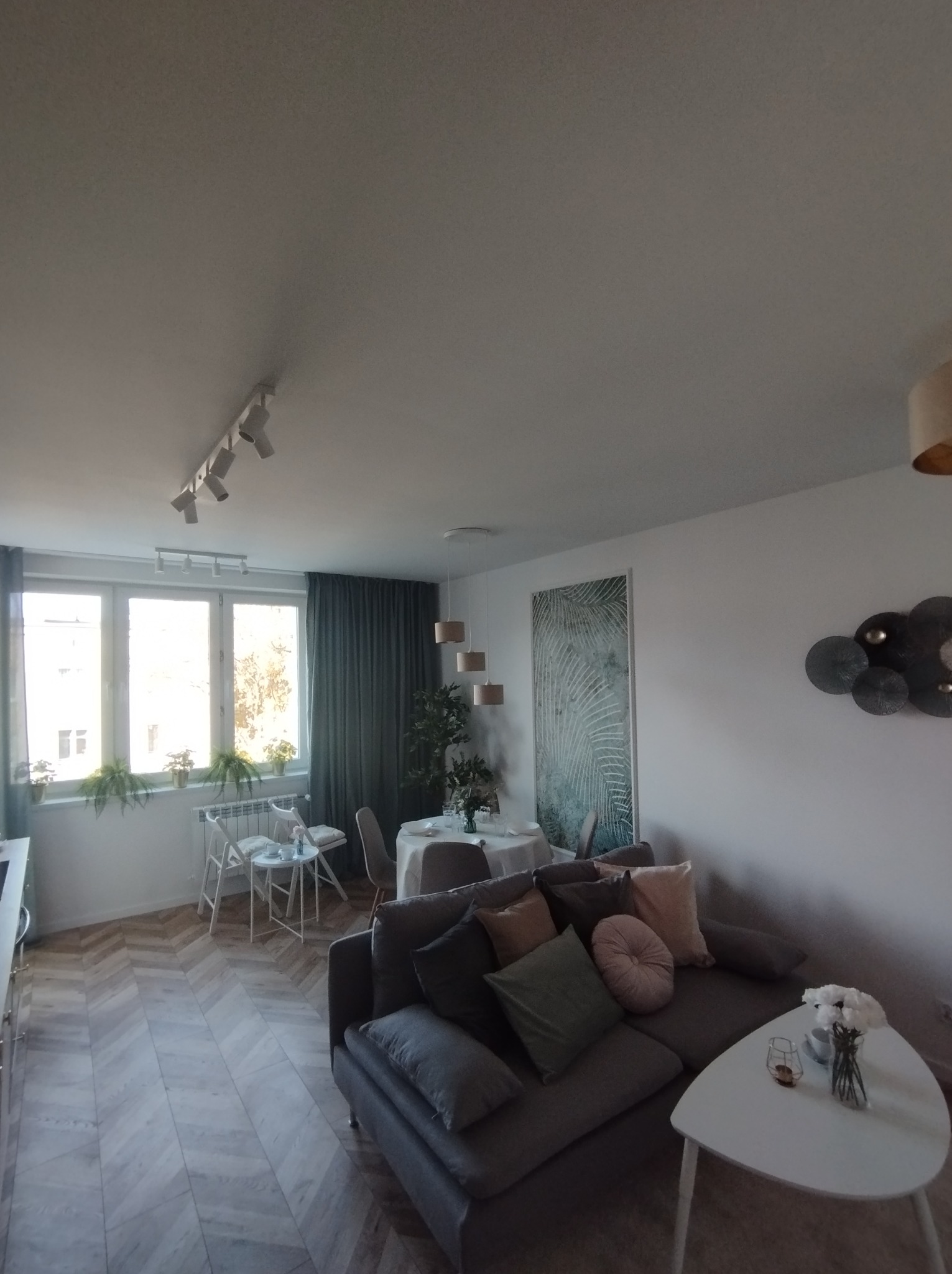 Jasne wnętrze salonu z parkietem ułożonym w jodełkę, szarą sofą z poduszkami, białym stolikiem kawowym i dekoracjami ściennymi.