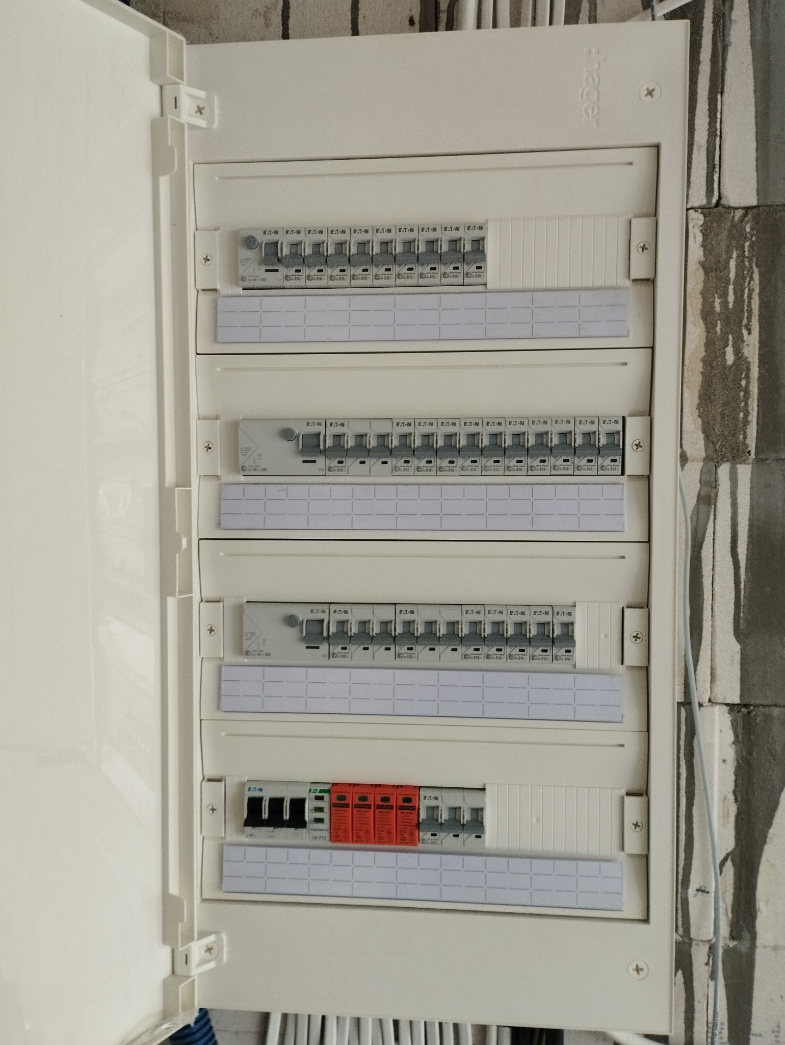 Otwarta skrzynka elektryczna z bezpiecznikami EATON i Hager, zamontowana na surowej ścianie z betonu komórkowego, widoczne przewody w dolnej części.