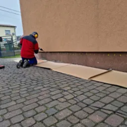 Pracownik w czerwonej kurtce wierci otwory w ścianie budynku, zabezpieczonej kartonem, prawdopodobnie pod iniekcję poziomą, z widocznymi zielonymi punktami wyznaczającymi miejsca wiercenia.