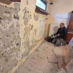 Pracownik wierci otwory w ścianie z widocznymi śladami wilgoci i ż&oacute;łtymi kołkami, przygotowując ją do iniekcji; widoczny młot udarowy i wiadro.