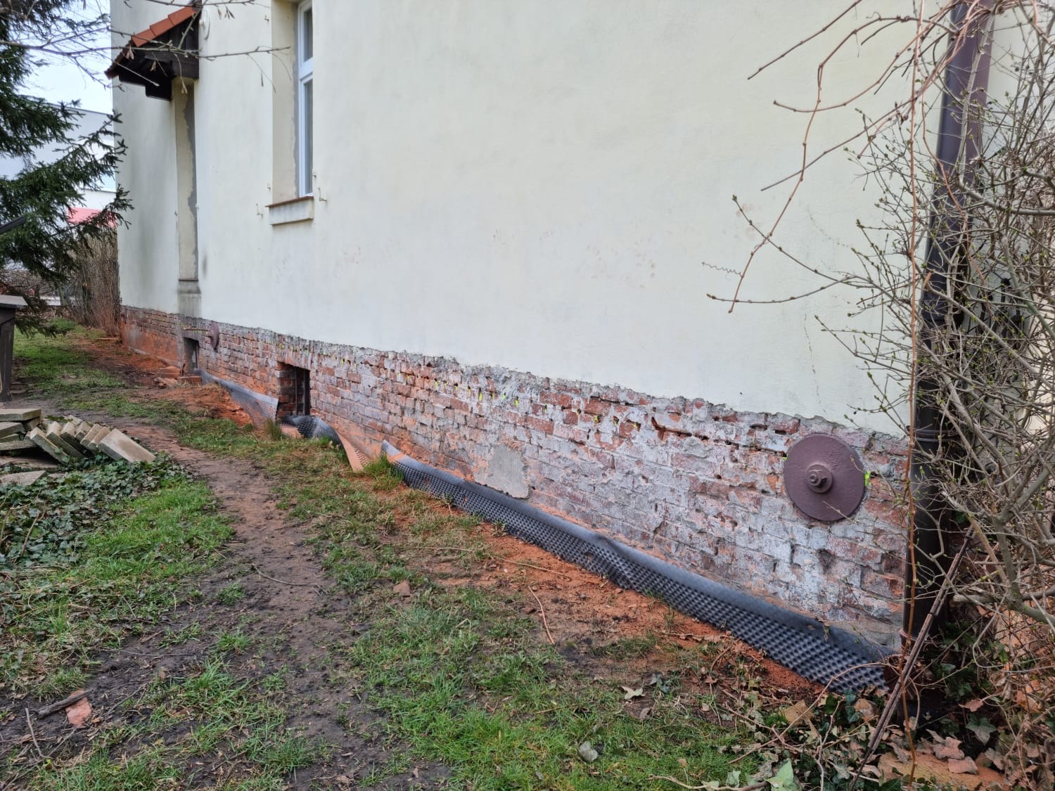 Przygotowanie do iniekcji poziomej w murze fundamentowym z odsłoniętą cegłą, widoczne wiercenia i membrana kubełkowa przy gruncie, na ścianie metalowa kotwa.