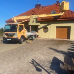 Żółty podnośnik koszowy z napisem 'Mycie Ciśnieniowe' ustawiony na kostce brukowej przed domem z czerwoną dachówką, podczas wykonywania prac na wysokości.