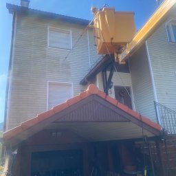 Mycie Ciśnieniowe Luiza Barska - Pracownik w kasku, w żółtym podnośniku koszowym, spryskuje wodą elewację domu z drewnianą okładziną, widoczne ślady czyszczenia.