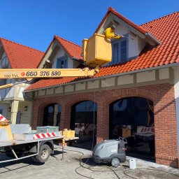 Mycie Ciśnieniowe Luiza Barska - Pracownik w żółtym kombinezonie w koszu podnośnika koszowego czyści elewację domu z czerwoną dachówką za pomocą myjki ciśnieniowej; na pierwszym planie widoczna myjka Kärcher i wąż ciśnieniowy...