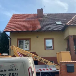 Częściowo wyczyszczony dach z czerwonej dachówki, widoczna różnica w kolorze, platforma podnośnika koszowego i częściowo widoczny samochód dostawczy z reklamą usług.