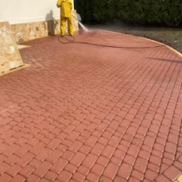 Pracownik w żółtym kombinezonie czyści czerwoną kostkę brukową na podjeździe za pomocą myjki ciśnieniowej, widoczne obłoki pary wodnej, obrzeże kostki w kolorze żółtym, dom z elewacją w tle.