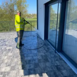 Pracownik w żółtej kamizelce i zielonych kaloszach czyści elewację domu wysokociśnieniową myjką na tarasie z kostki brukowej. W tle widoczne drzewa i zabudowania.