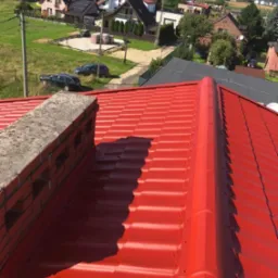 Czerwony dach z blachy trapezowej po renowacji, widok z góry na komin z cegły i okoliczne domy w słoneczny dzień.