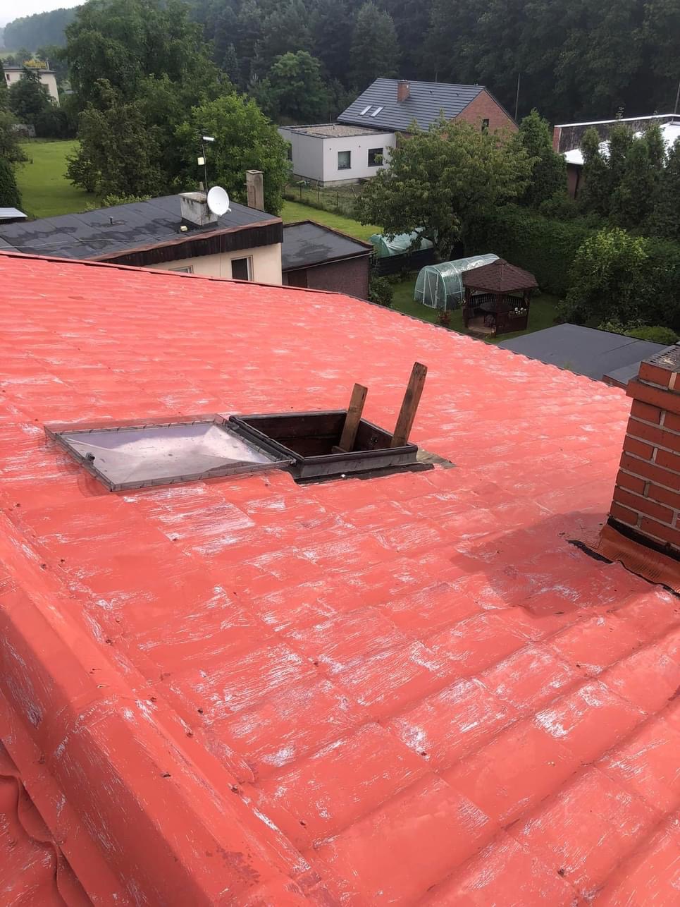 Czerwony dach z otwartym oknem dachowym i widocznymi śladami świeżego gruntowania, w tle zabudowa jednorodzinna i zieleń