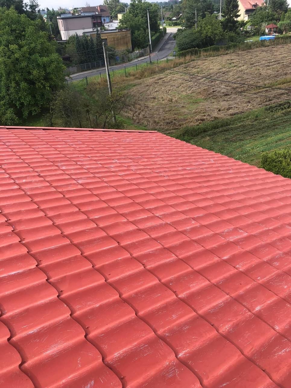 Czerwona dachówka ceramiczna po renowacji, widok z góry na tle zieleni i zabudowań mieszkalnych w oddali.