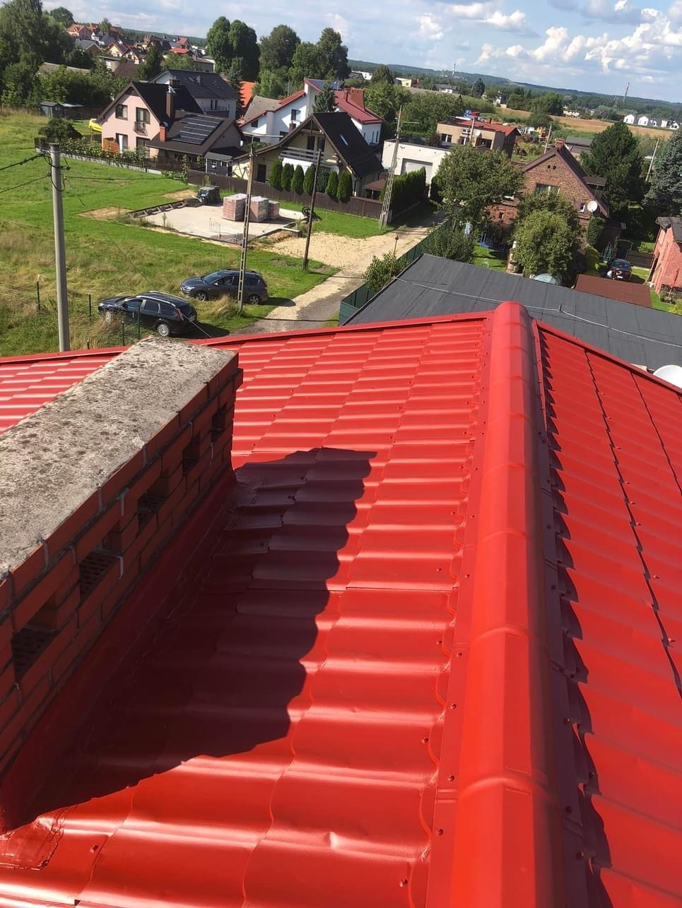Czerwony dach z blachy trapezowej po renowacji, widok z góry na komin z cegły i okoliczne domy w słoneczny dzień.