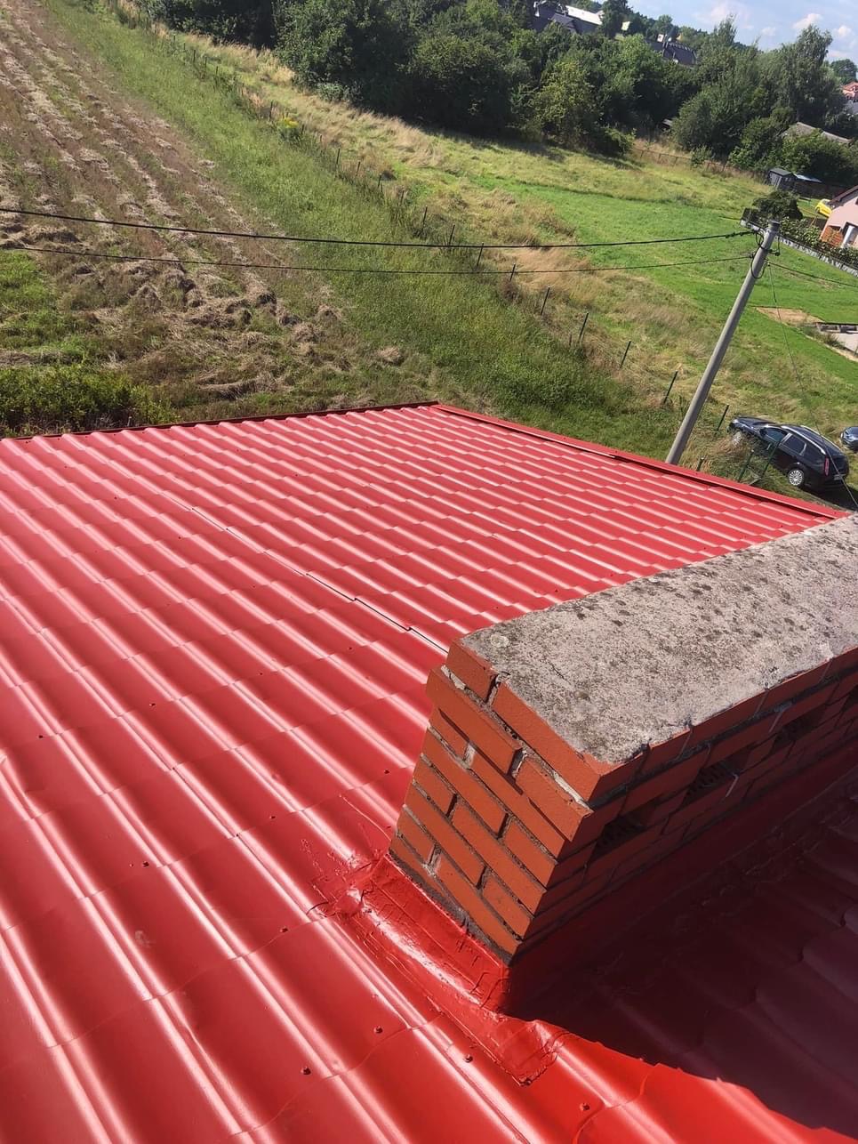 Czerwony blaszany dach po renowacji, z uszczelnionym kominem z cegły, na tle zielonego pola i zabudowań.