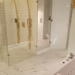 Luksusowa łazienka z kabiną prysznicową walk-in, złotymi deszczownicami i armaturą, białą marmurową podłogą i ścianami z dekoracyjnymi złotymi detalami, oraz widoczną częścią wanny z hydromasażem.