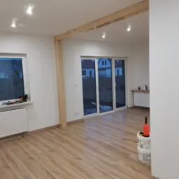 Jasne wnętrze w trakcie wykańczania, z drewnianą konstrukcją stropu, świeżo położoną podłogą laminowaną, widocznymi narzędziami malarskimi i pojemnikami po farbie.