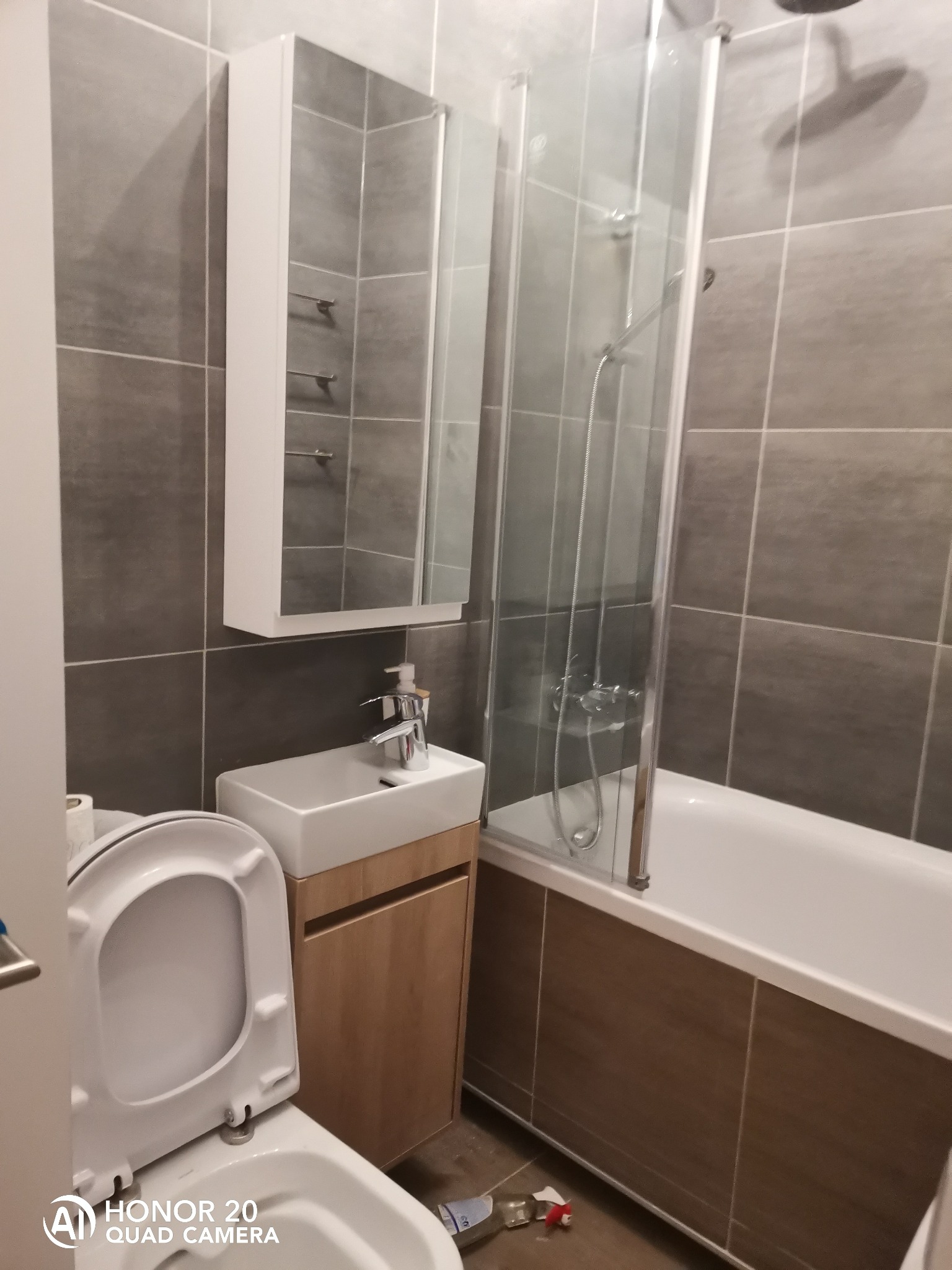 Niewielka łazienka z szarymi płytkami, białą umywalką nablatową na drewnianej szafce, lustrzaną szafką wiszącą i kabiną prysznicową nad wanną.