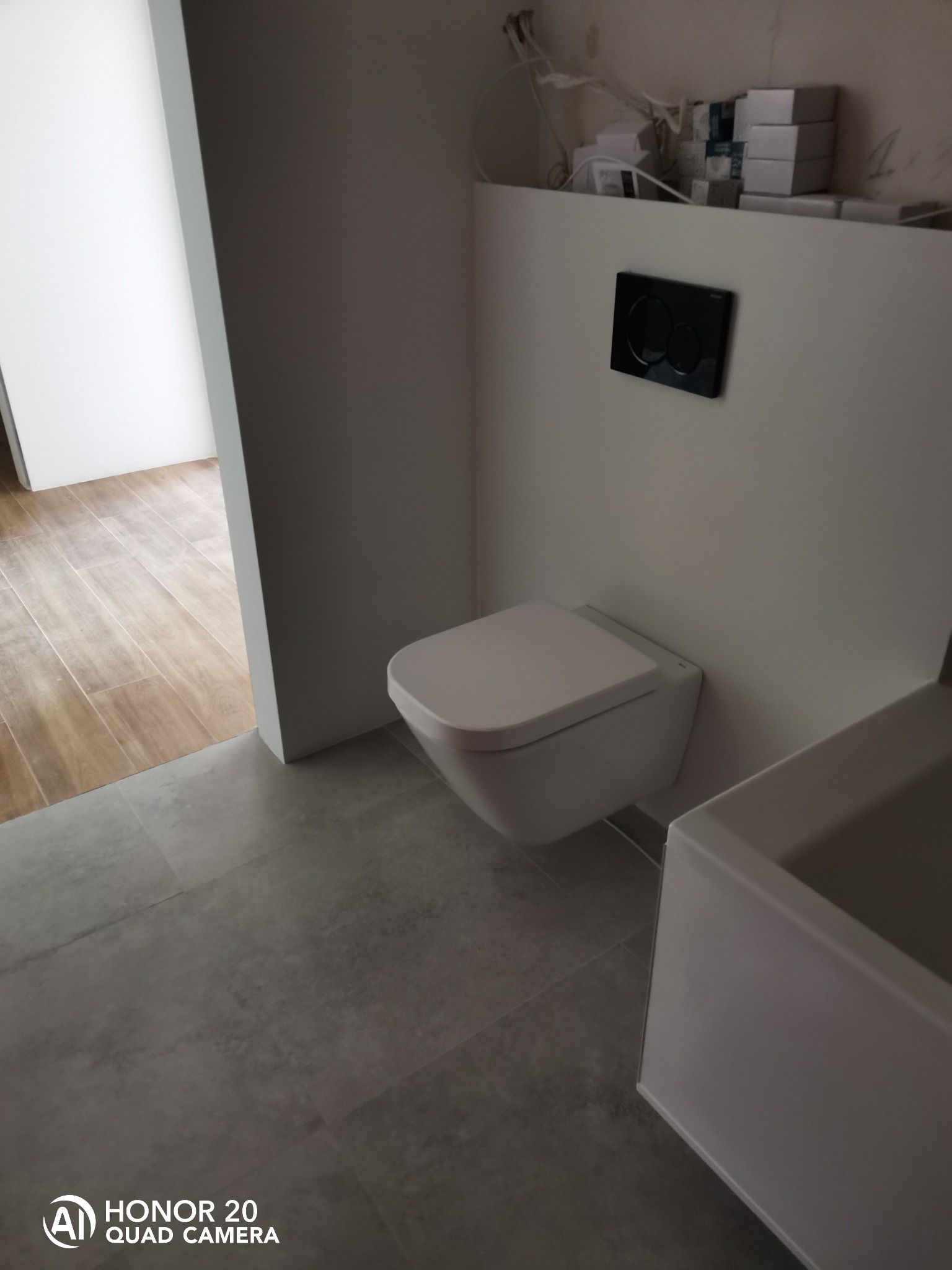 Nowoczesna toaleta podwieszana z czarnym przyciskiem spłukującym na białej ścianie, szare płytki podłogowe, widoczne instalacje na górze ściany, fragment umywalki i podłoga z drewnopodobnych płytek...