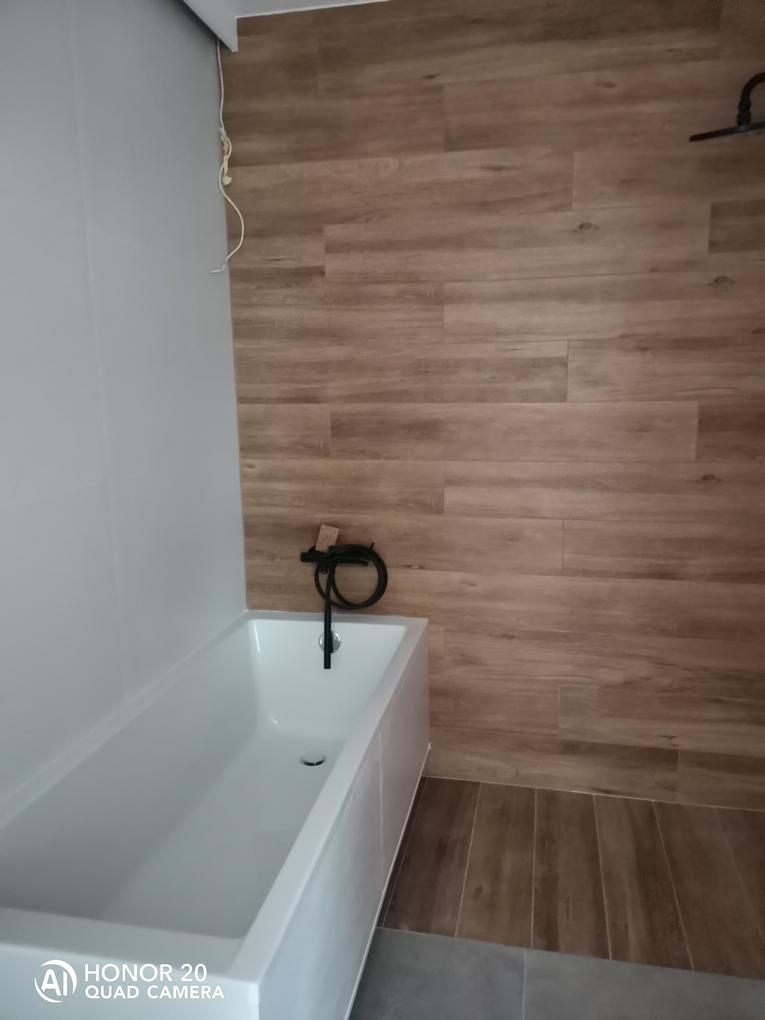 Biała wanna z czarną armaturą na tle ściany wyłożonej brązowymi płytkami imitującymi drewno, z widocznym fragmentem szarej podłogi i wystającym kablem elektrycznym.
