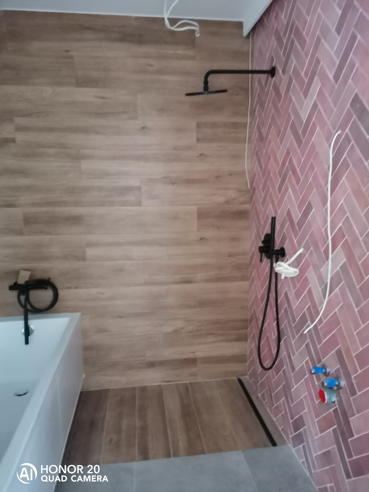 Łazienka w trakcie remontu: różowe płytki ułożone w jodełkę na ścianie, drewnopodobne płytki na ścianie, czarna armatura, widoczne przewody i instalacje wodne.