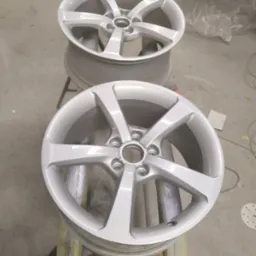 Dwa odnowione aluminiowe felgi samochodowe w srebrnym kolorze, ustawione jeden na drugim na metalowych stojakach w warsztacie, widoczne ślady lakierowania.