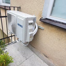 Makowski Group Energy - Biały klimatyzator HEIKO zamontowany na balkonie, widoczny z boku, z fragmentem balustrady i rośliny. Elewacja budynku w tle, okno z ciemnym parapetem.