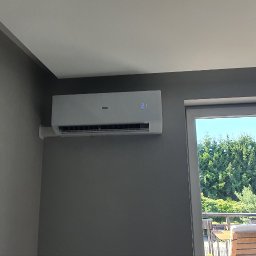 Makowski Group Energy - Biały klimatyzator Haier z wyświetlaczem temperatury (21°C) zamontowany na szarej ścianie w pokoju z widokiem na zieleń przez okno balkonowe.