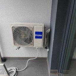 Makowski Group Energy - Klimatyzator Haier zamontowany na balkonie z widokiem na zieleń miejską. Biała obudowa, niebieskie logo, szare płytki podłogowe i częściowo widoczne krzesło.