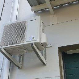 Makowski Group Energy - Biały klimatyzator marki Innova zamontowany na wspornikach na elewacji budynku z widoczną rurą odprowadzającą skropliny i fragmentem bramy garażowej.