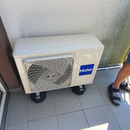 Makowski Group Energy - Biała jednostka zewnętrzna klimatyzacji Haier na balkonie, zamontowana na czarnych podkładkach antywibracyjnych. Widoczny fragment osoby w szortach i sandałach.