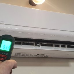 Makowski Group Energy - Biała klimatyzacja Mitsubishi Heavy Industries na ścianie, mierzona termometrem. Widoczny wskaźnik temperatury 51.3. Oświetlenie sufitowe i fragment lampki w tle.