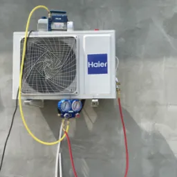 Biała jednostka zewnętrzna klimatyzacji Haier zamontowana na szarej ścianie, podłączona do manometrów i pompy próżniowej podczas serwisu.