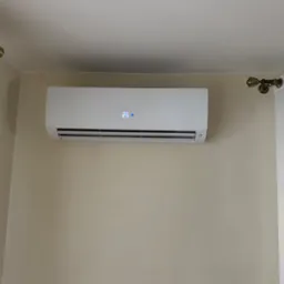 Biała klimatyzacja ścienna marki Haier z wyświetlaczem temperatury 25 stopni, zamontowana na beżowej ścianie w pomieszczeniu z białymi zasłonami.