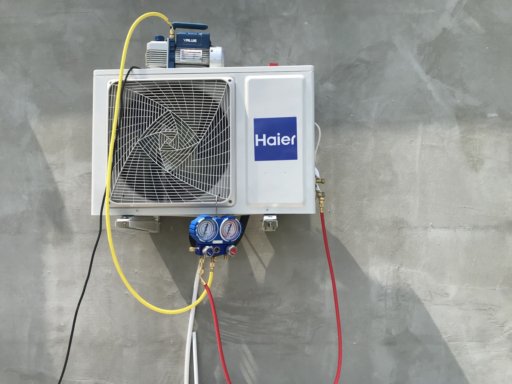 Biała jednostka zewnętrzna klimatyzacji Haier zamontowana na szarej ścianie, podłączona do manometrów i pompy próżniowej podczas serwisu.