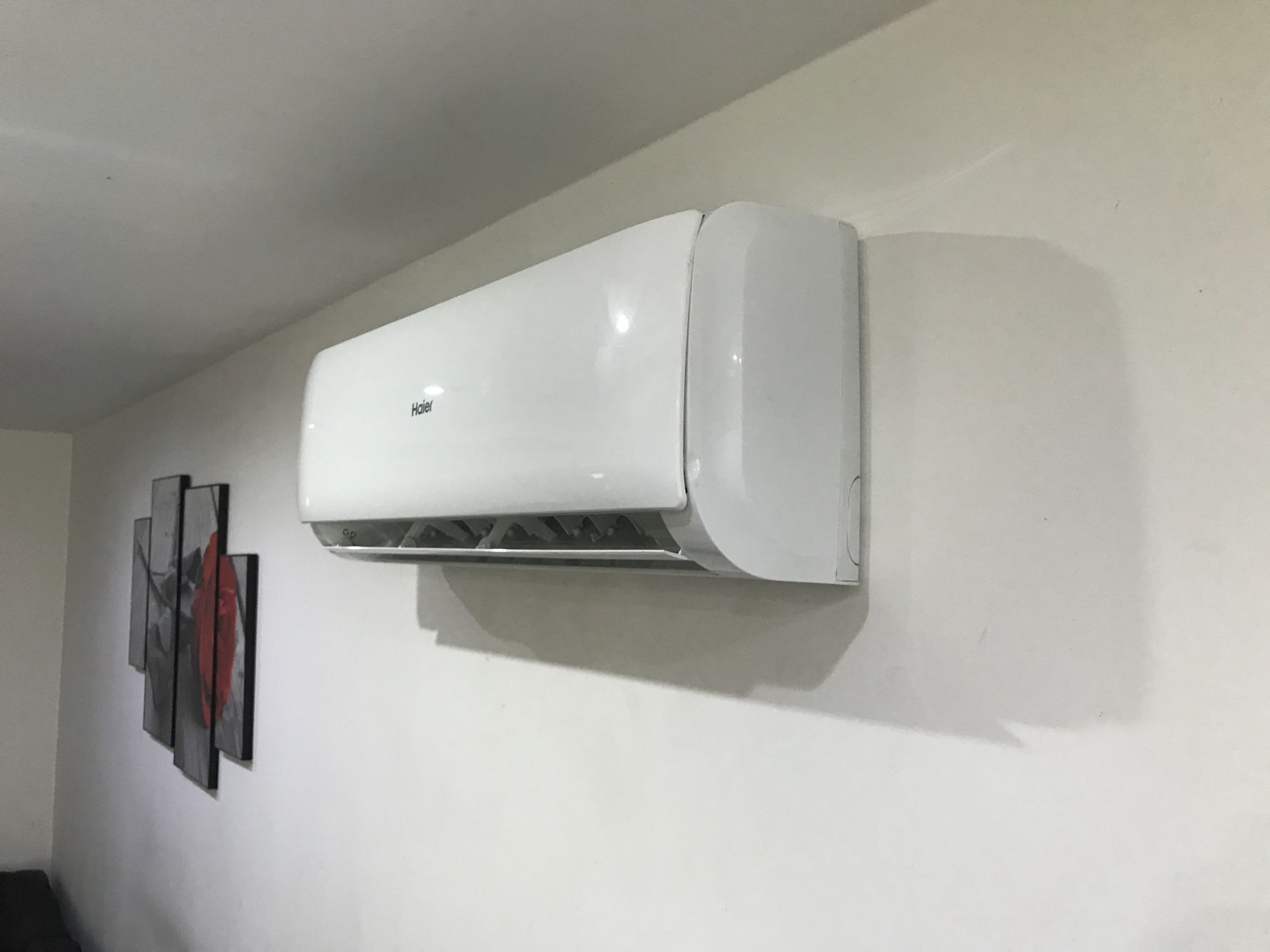 Biała jednostka wewnętrzna klimatyzatora Haier zamontowana na ścianie w pomieszczeniu. Na ścianie obok wisi obraz złożony z kilku części w odcieniach szarości z czerwonym akcentem.