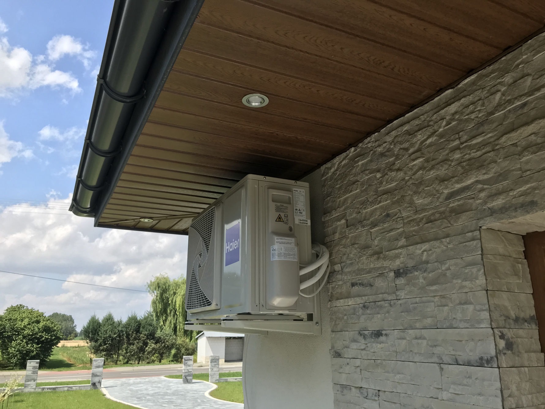 Zewnętrzna jednostka klimatyzacji Haier zamontowana na elewacji z kamienia dekoracyjnego, pod drewnianą podbitką dachu, widok na ogród i błękitne niebo z chmurami.