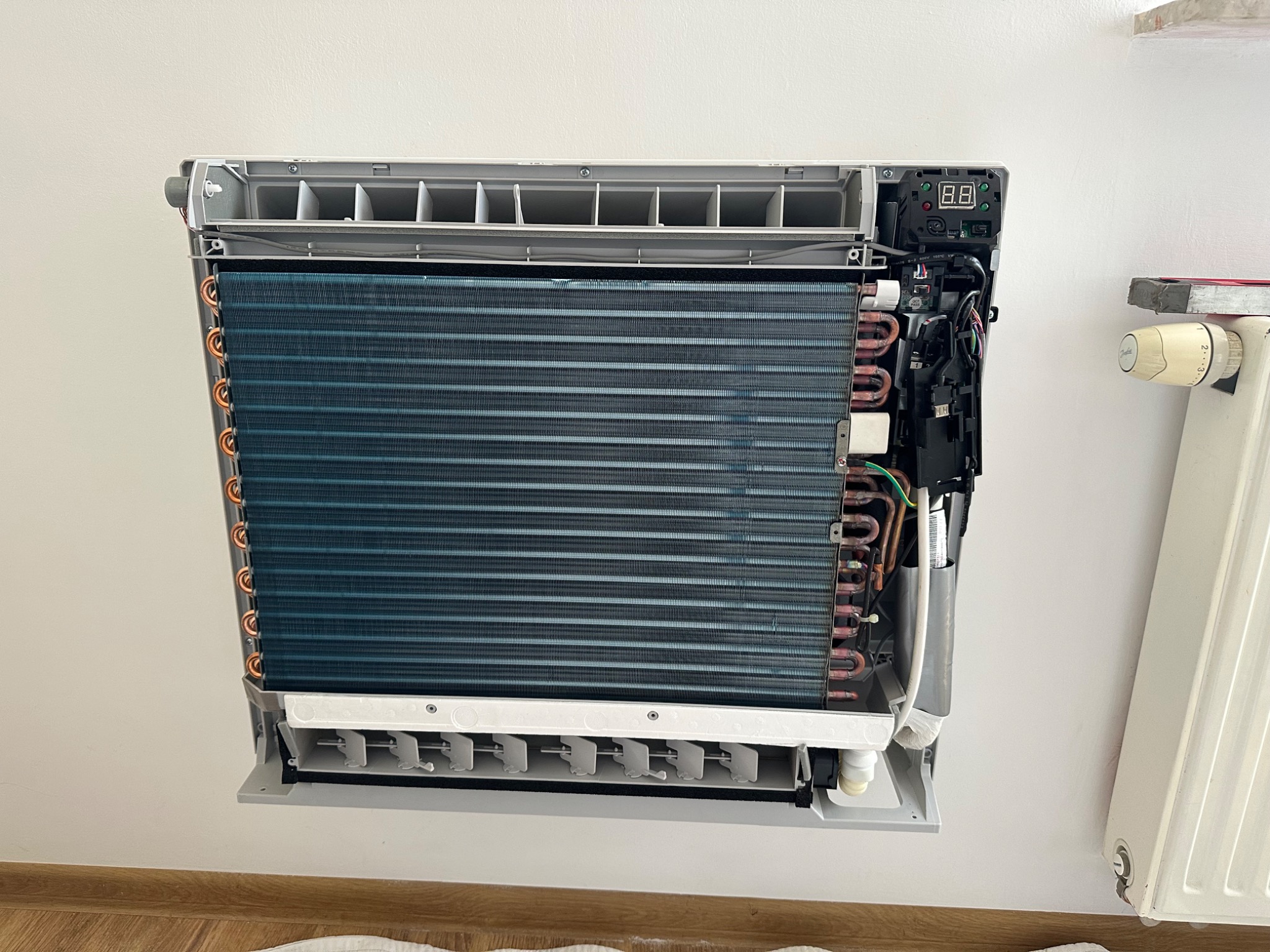 Wnętrze ściennego klimatyzatora z widocznymi elementami chłodzącymi i elektroniką, zamontowanego na białej ścianie obok grzejnika.