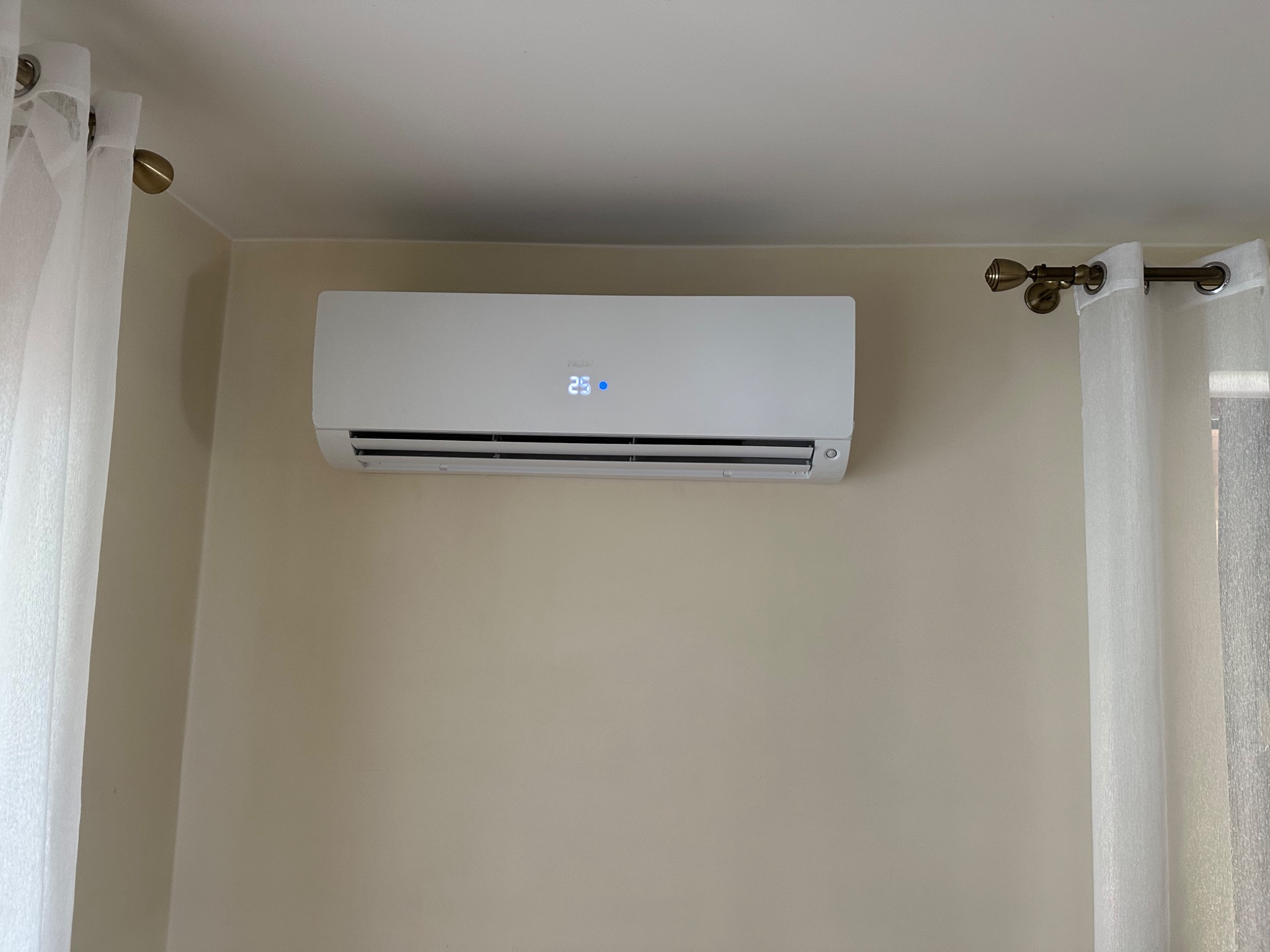 Biała klimatyzacja ścienna marki Haier z wyświetlaczem temperatury 25 stopni, zamontowana na beżowej ścianie w pomieszczeniu z białymi zasłonami.