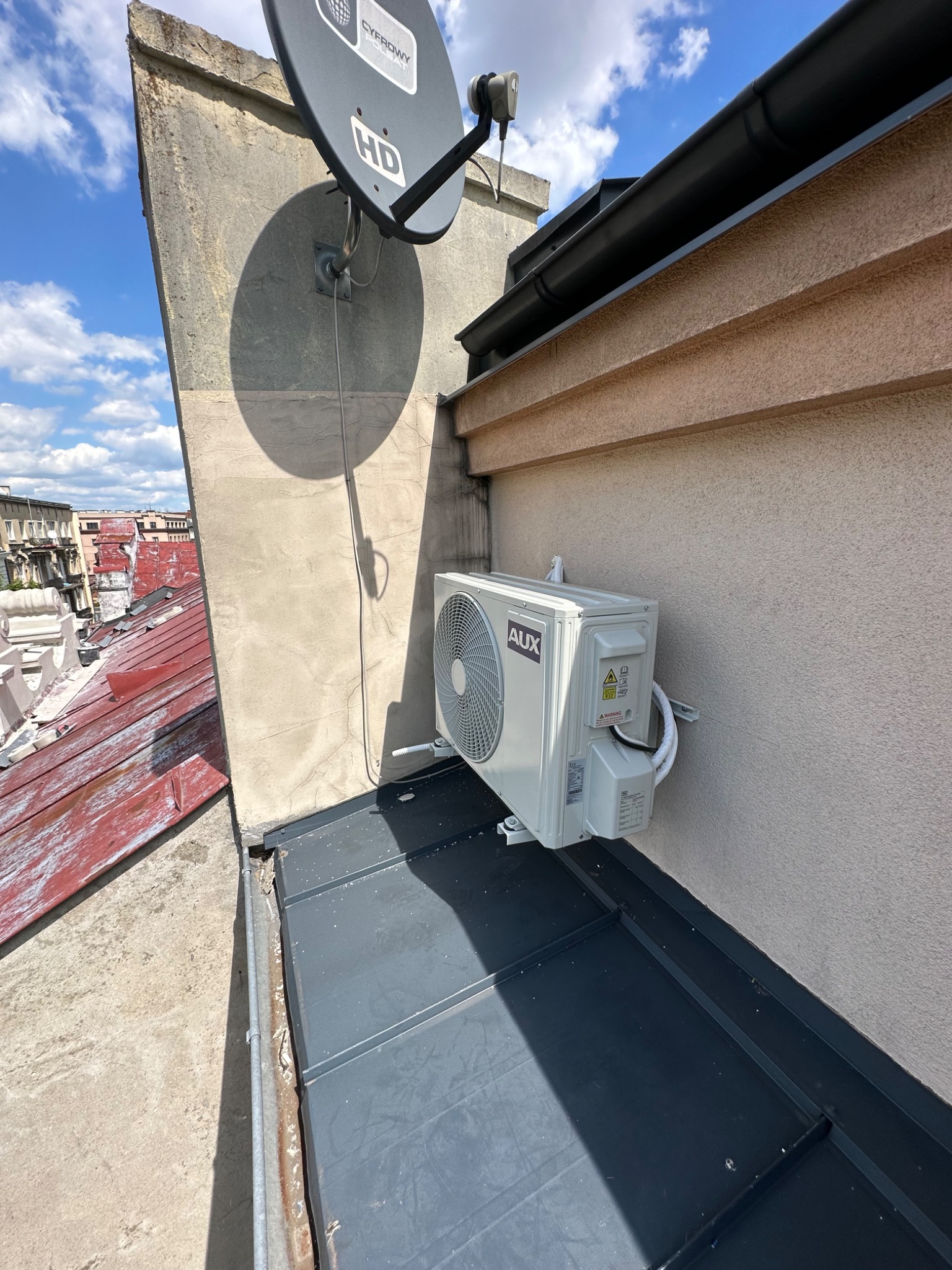 Zewnętrzna jednostka klimatyzacji marki AUX zamontowana na dachu budynku, z widoczną anteną satelitarną i fragmentem czerwonego dachu w tle.