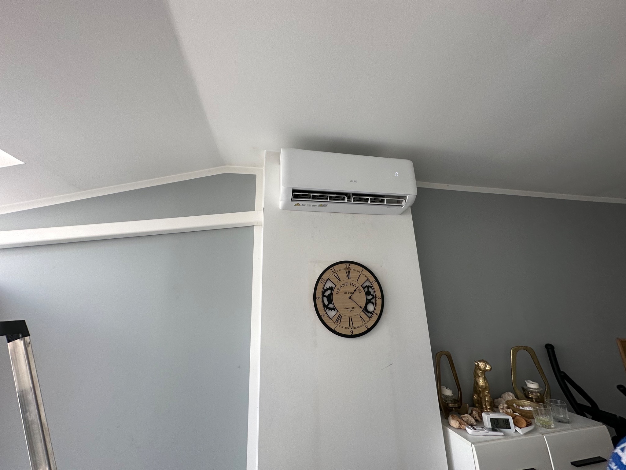 Biała jednostka wewnętrzna klimatyzacji zamontowana na ścianie w pomieszczeniu z zegarem w stylu retro i elementami dekoracyjnymi na komodzie. Widoczna drabina w rogu.