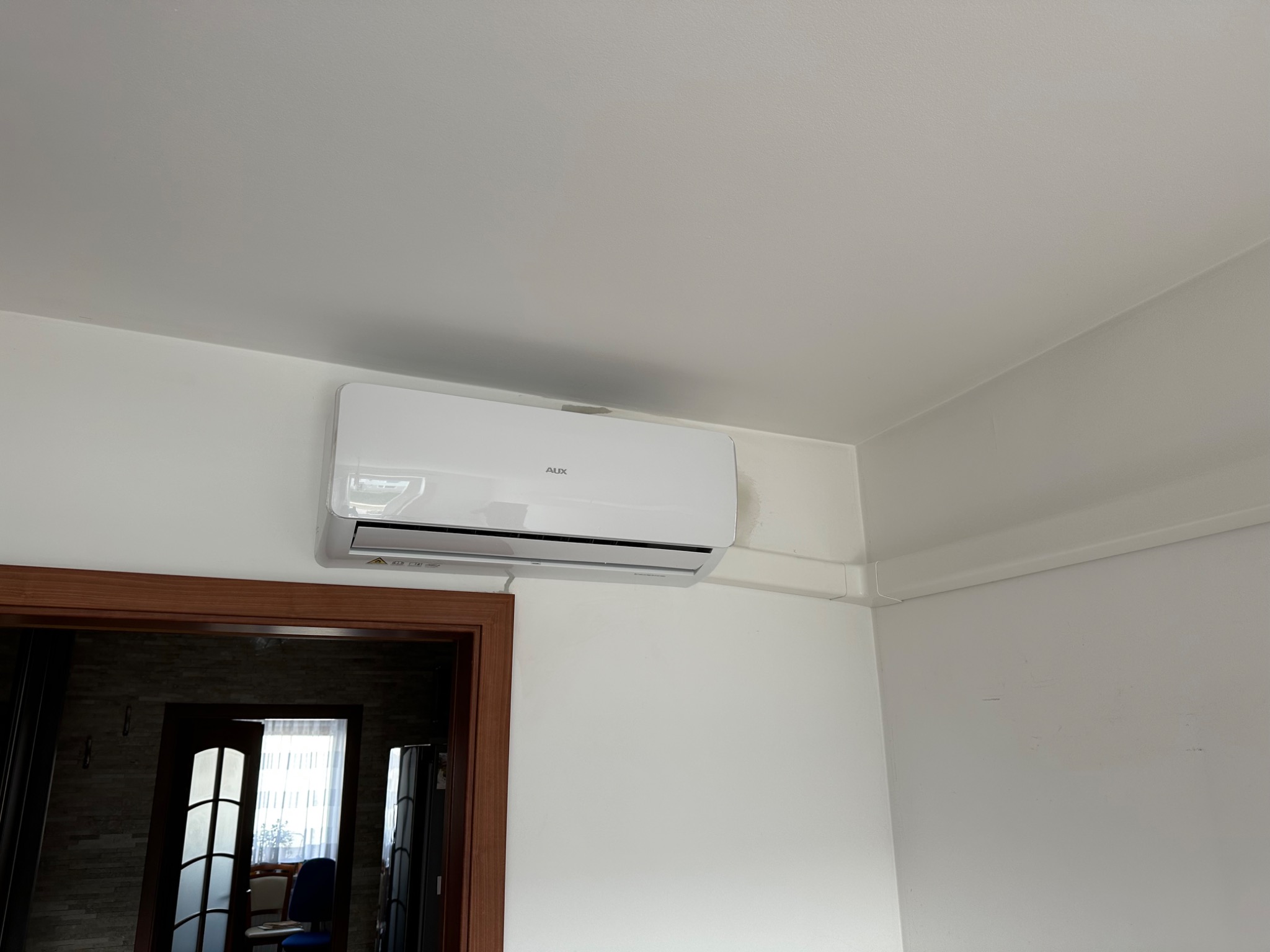 Biała klimatyzacja AUX zamontowana na ścianie w pomieszczeniu, widoczne białe listwy maskujące przewody, w tle otwarte drzwi do innego pokoju.
