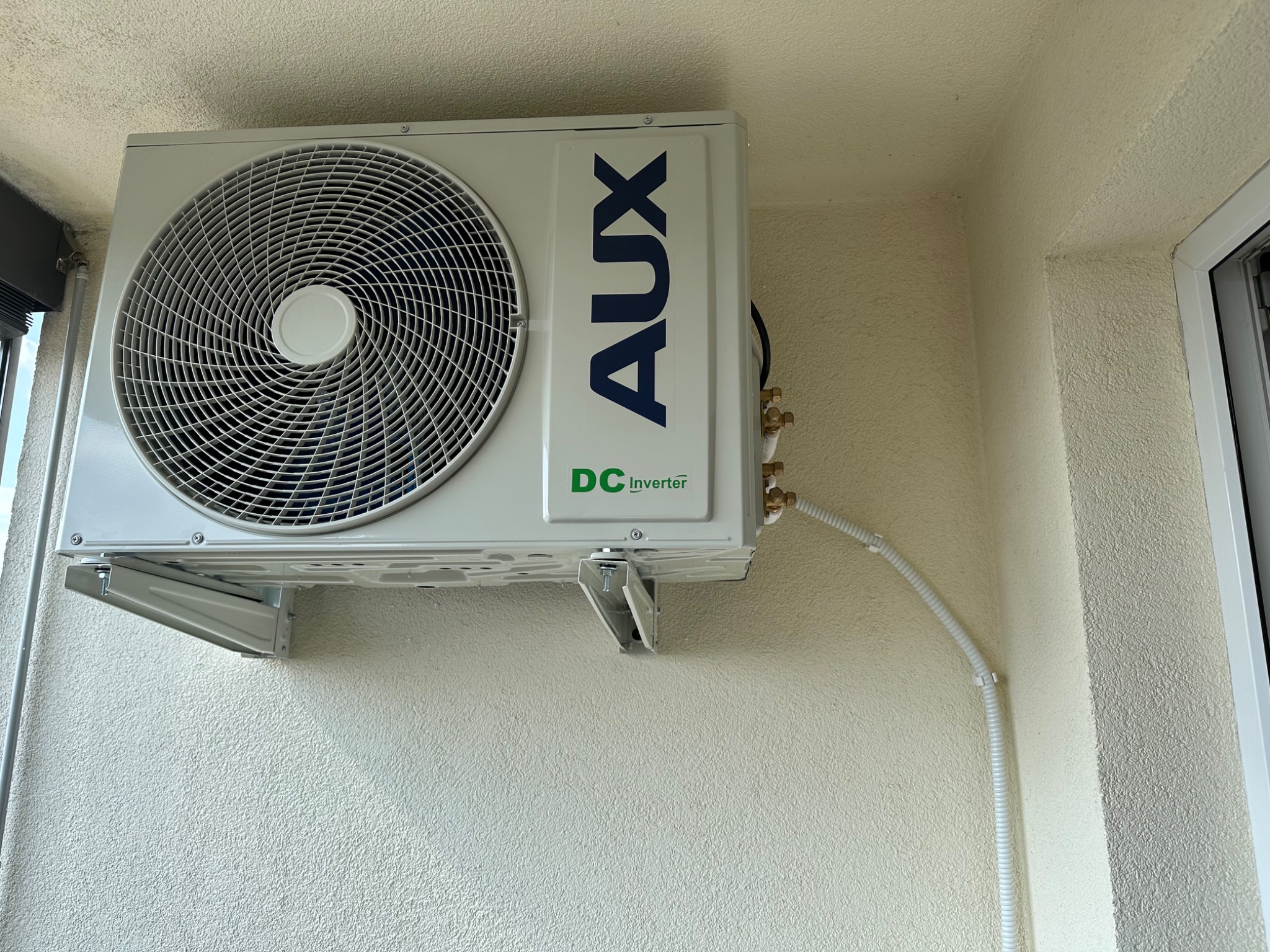 Zewnętrzna jednostka klimatyzacji AUX z widocznymi przyłączami i kablem w białej osłonie, zamontowana na jasnej, chropowatej ścianie zewnętrznej budynku.