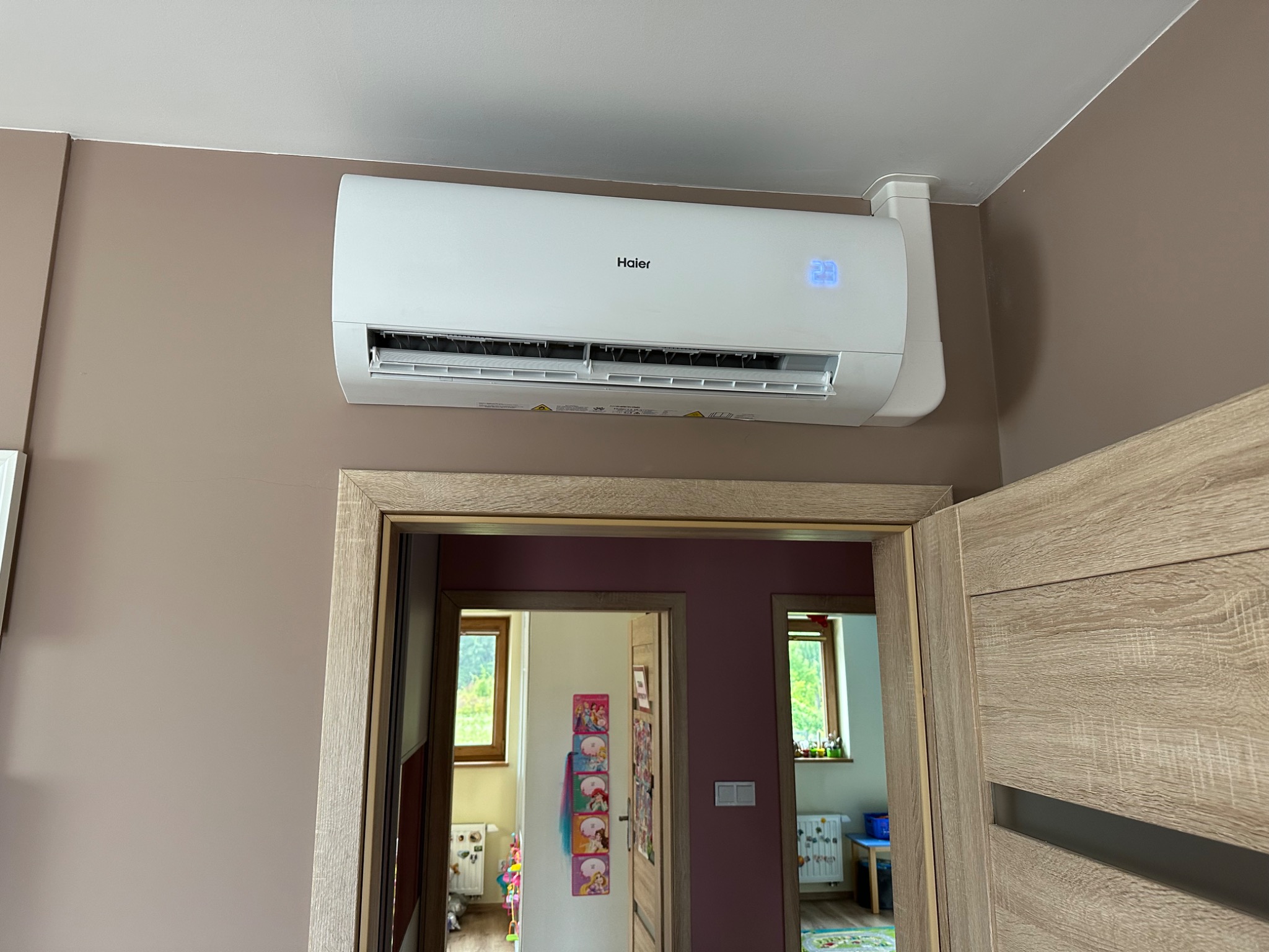 Biały klimatyzator Haier zamontowany na brązowej ścianie nad drewnianą ościeżnicą drzwi, widok na dwa pokoje dziecięce z oknami.