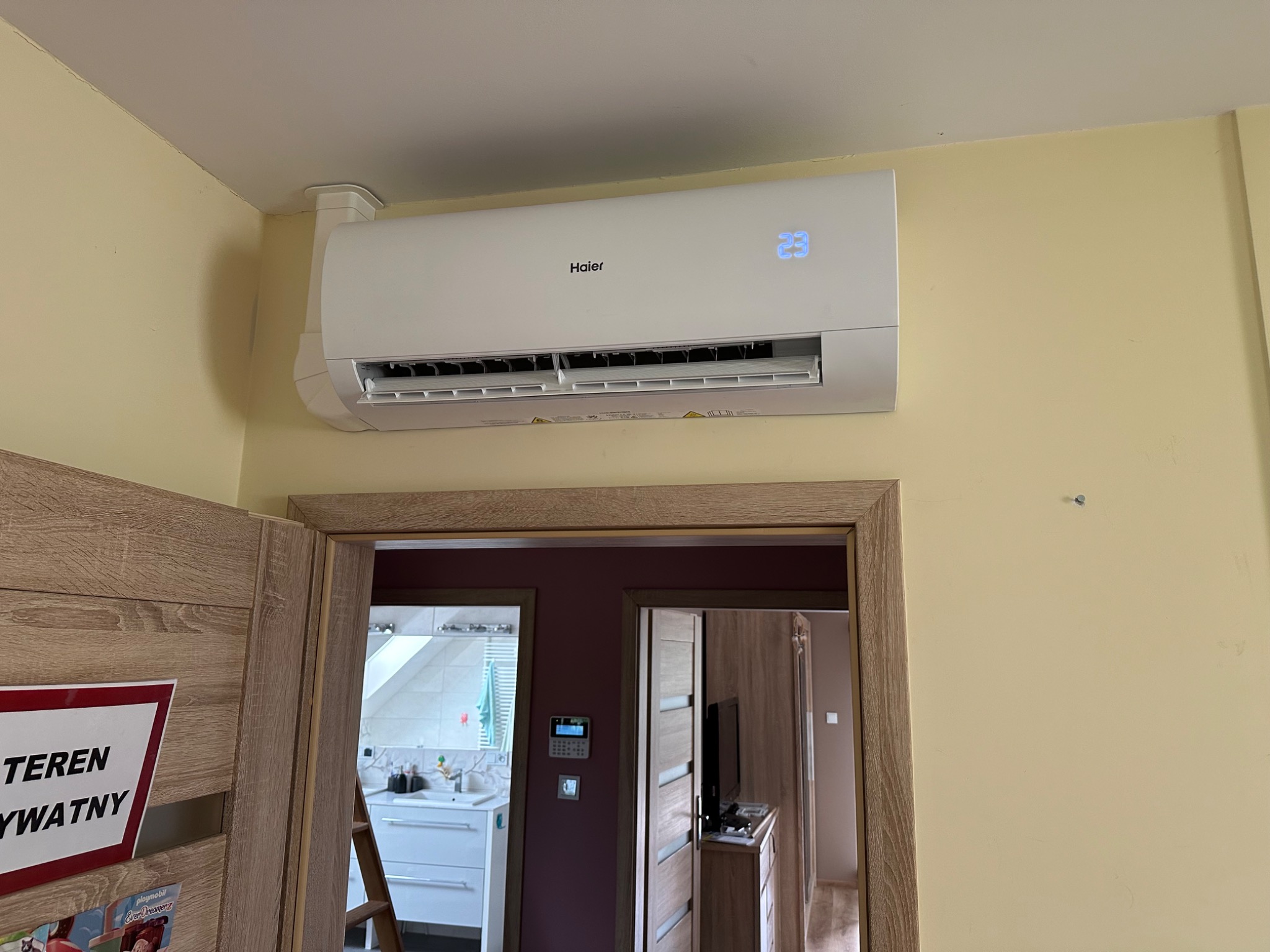 Biały klimatyzator Haier zamontowany nad drzwiami w żółtym pokoju, widok na sąsiednie pomieszczenia przez otwarte drzwi.