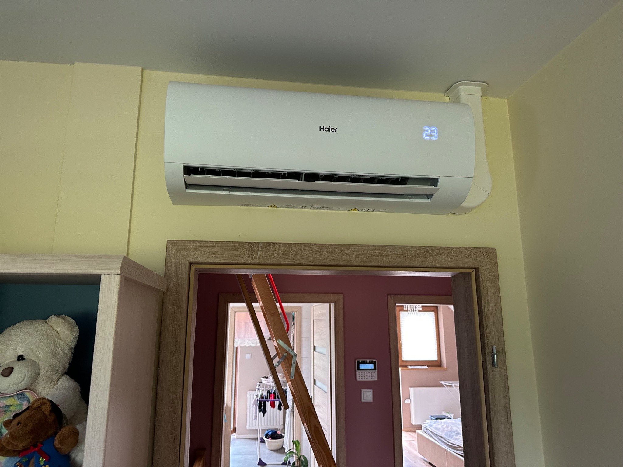 Biały klimatyzator Haier zamontowany na żółtej ścianie nad drewnianą futryną drzwi, widoczny wyświetlacz z temperaturą 23 stopnie Celsjusza, przez otwarte drzwi widoczny pokój z suszarką na pranie...