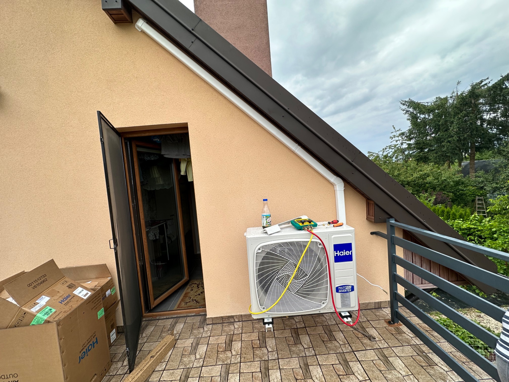 Zewnętrzna jednostka klimatyzacji Haier zamontowana na balkonie budynku, podłączona do manometrów serwisowych, obok otwarte drzwi balkonowe i kartony po klimatyzacji.