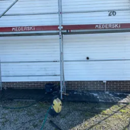 Biała elewacja domu z sidingu, rusztowanie z nazwą firmy 'MEDERSKI', myjka ciśnieniowa i mokry chodnik z kostki brukowej.