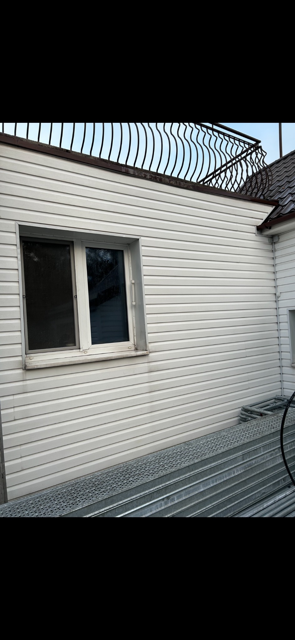Boczny widok budynku z białą elewacją z sidingu, widoczne okno z zabrudzeniami, metalowe rusztowanie złożone obok ściany i ozdobna metalowa balustrada na dachu.
