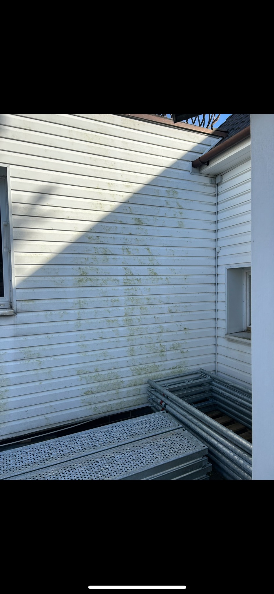 Biała elewacja sidingowa z widocznymi śladami zabrudzeń biologicznych i stosem rusztowań na dole kadru.