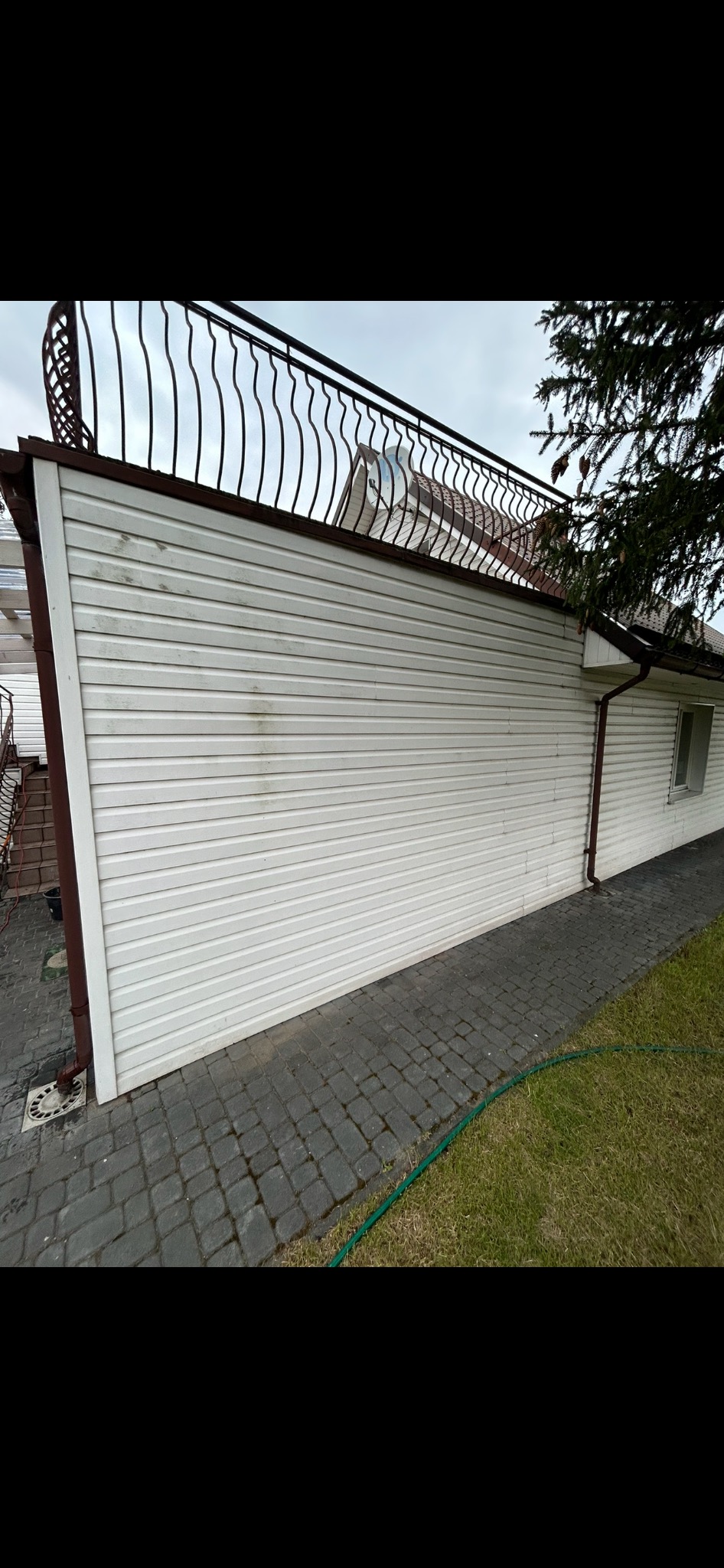 Boczny widok elewacji domu z sidingu z widocznymi zabrudzeniami i ciemnymi plamami, z brązowym wykończeniem i ozdobną balustradą na dachu.