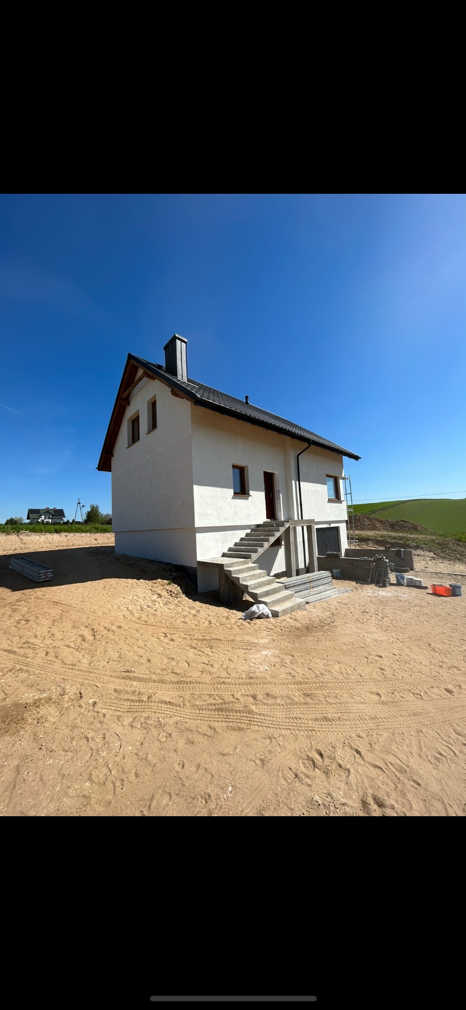 Biały dom jednorodzinny z szarymi schodami wejściowymi, świeżo po tynkowaniu elewacji, na piaszczystym terenie, z widocznymi śladami opon i odległym budynkiem w tle.