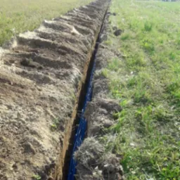 Długi wykop w ziemi z widocznymi niebieskimi rurami na dnie, świeżo usypana ziemia po bokach, dom w tle.